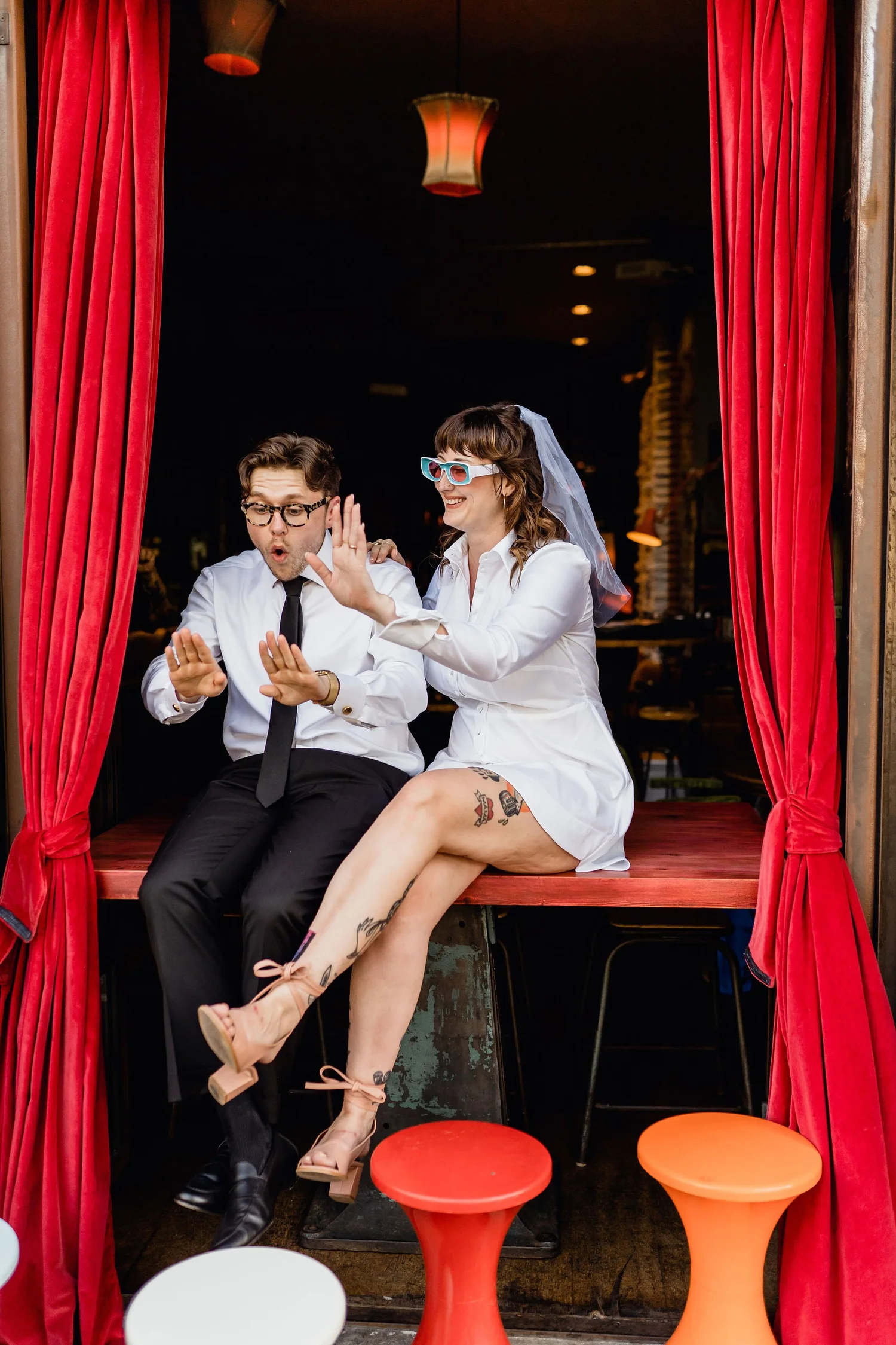 Two people wearing wedding attire sitting on a stage with red curtains, playful expressions, and colorful stools at their feet.