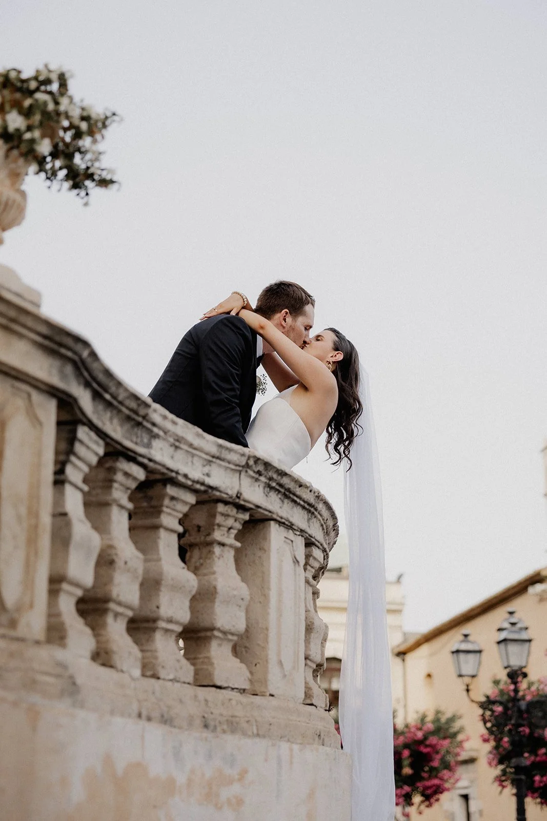 A couple dressed in wedding attire sharing a kiss on a stone balcony with a village and flowering trees in the background.
