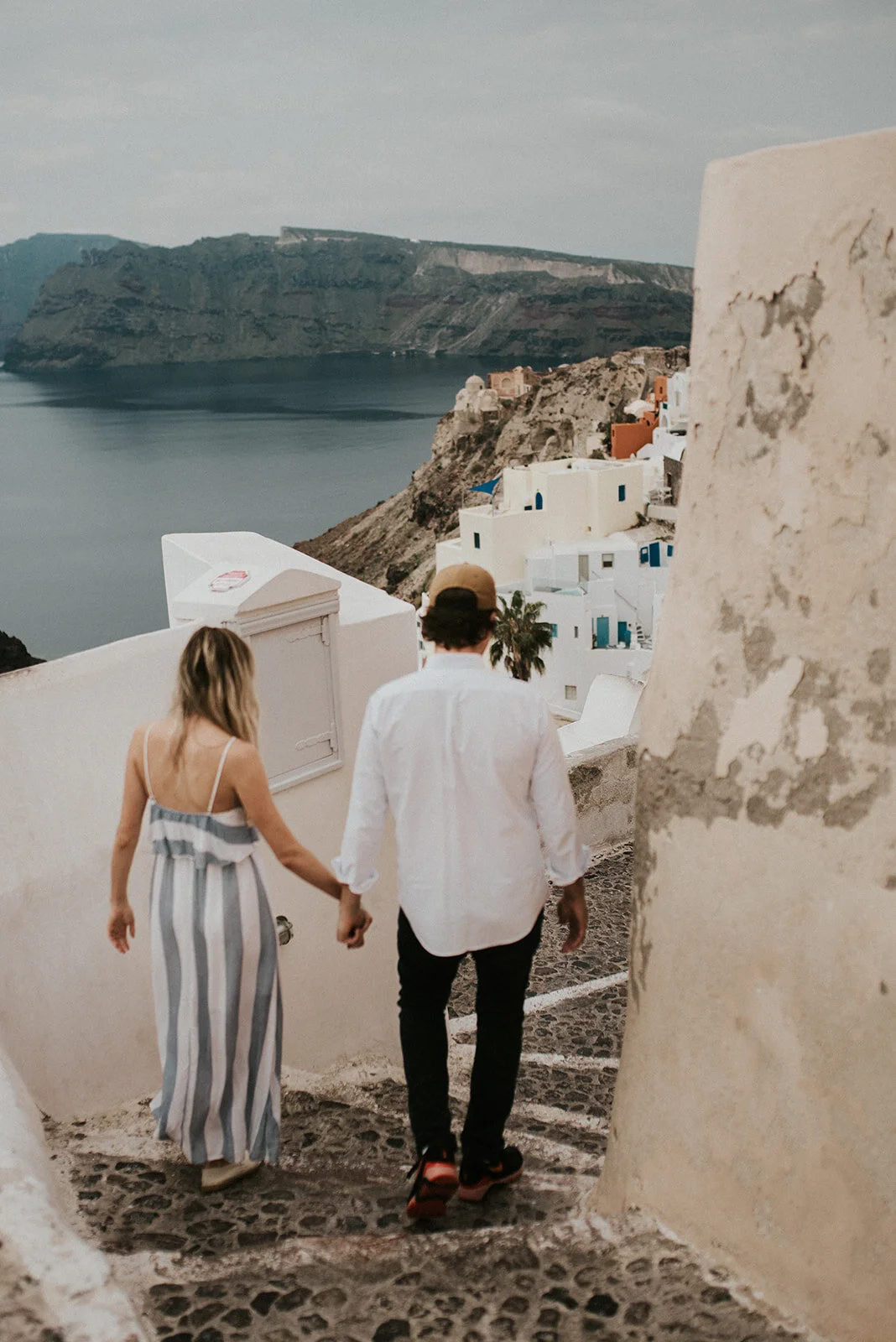 A couple holding hands walking down a stone pathway in a scenic coastal village with white buildings and cliffs in the background.