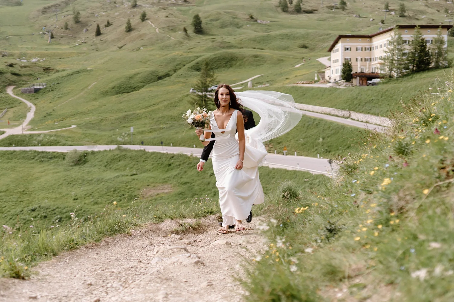 A bride in a white wedding dress holding a bouquet of flowers walking on a dirt path in a green hilly landscape with a large building in the background.