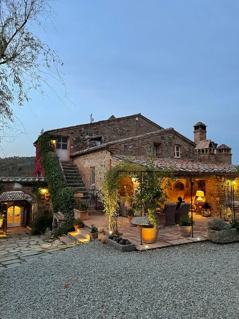 A rustic stone house with an outdoor patio, illuminated by warm lights, surrounded by potted plants and a gravel yard, with a tree on the left and a blue sky overhead.
