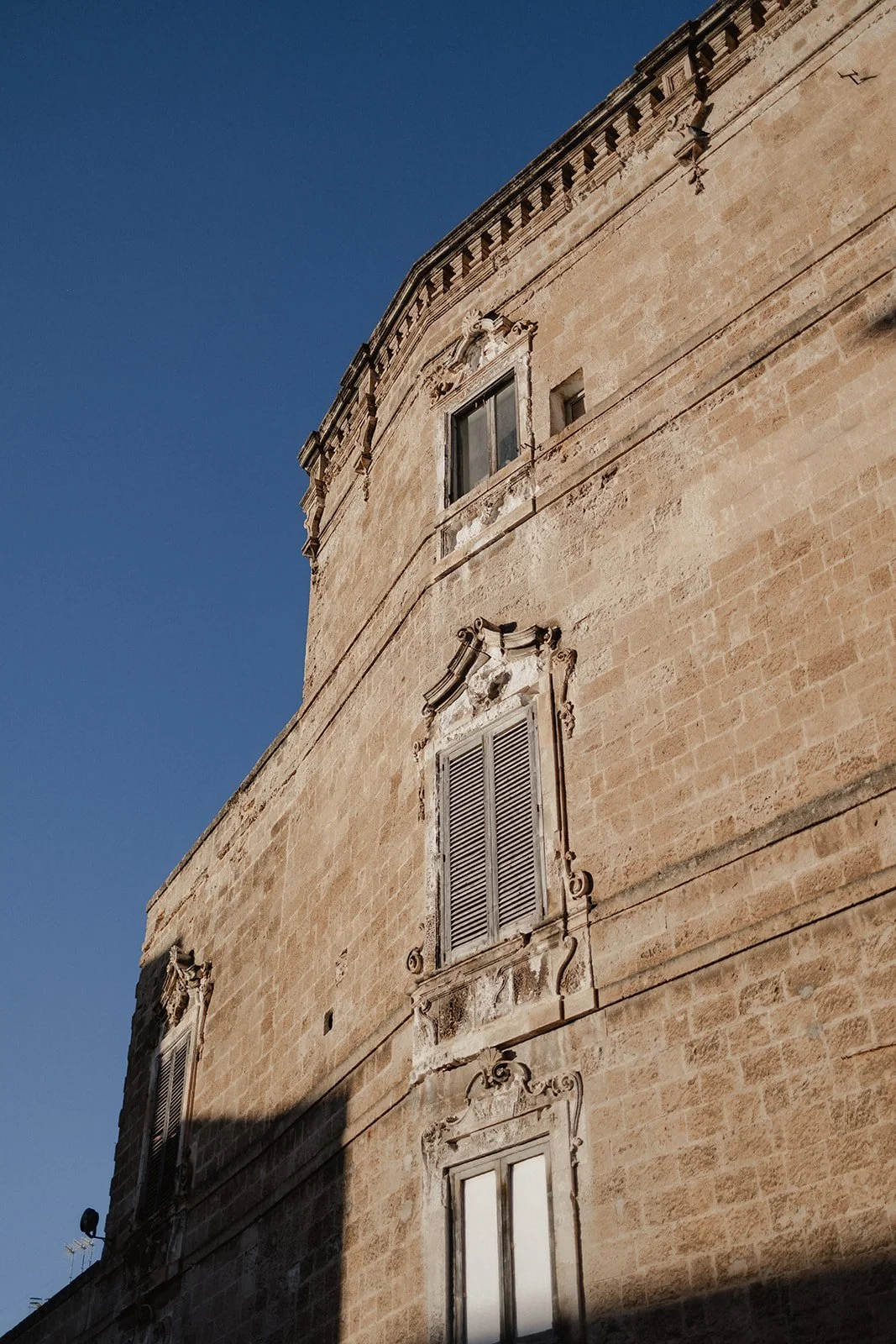 Close-up view of a historic building with beige stone facade, ornate window frames, and shutters against a clear blue sky.