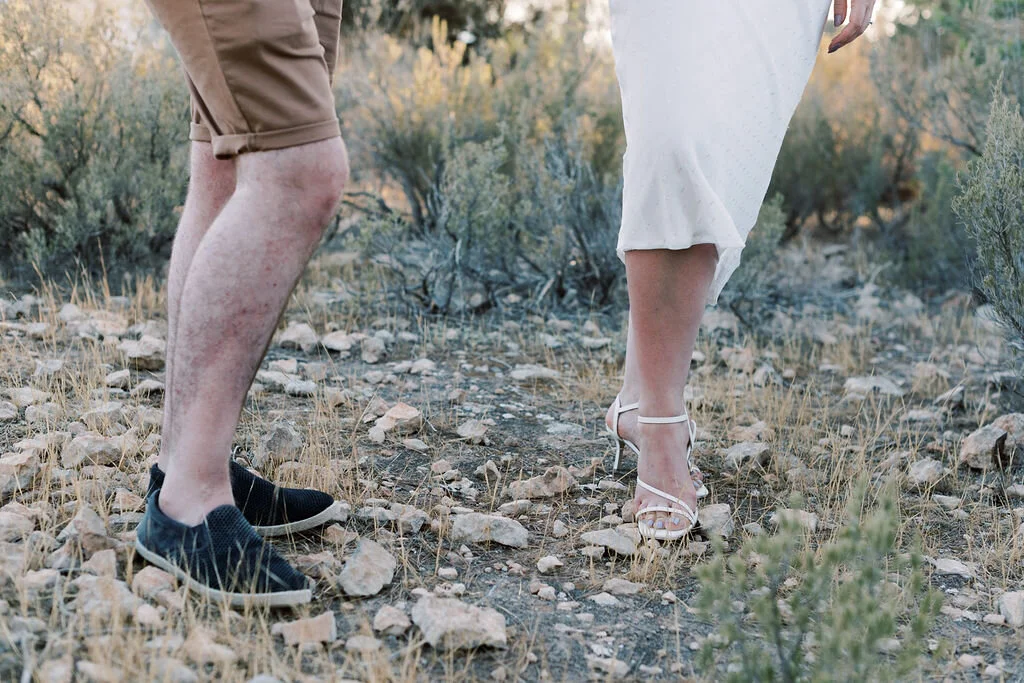Close-up of a man and woman standing outdoors on a rocky, dirt trail with sparse bushes.