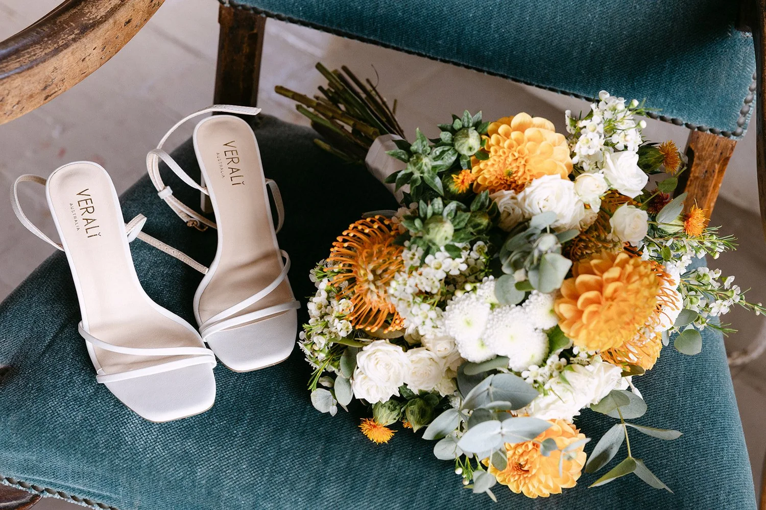 A pair of white high-heeled shoes and a bouquet of white and orange flowers placed on a blue upholstered chair.