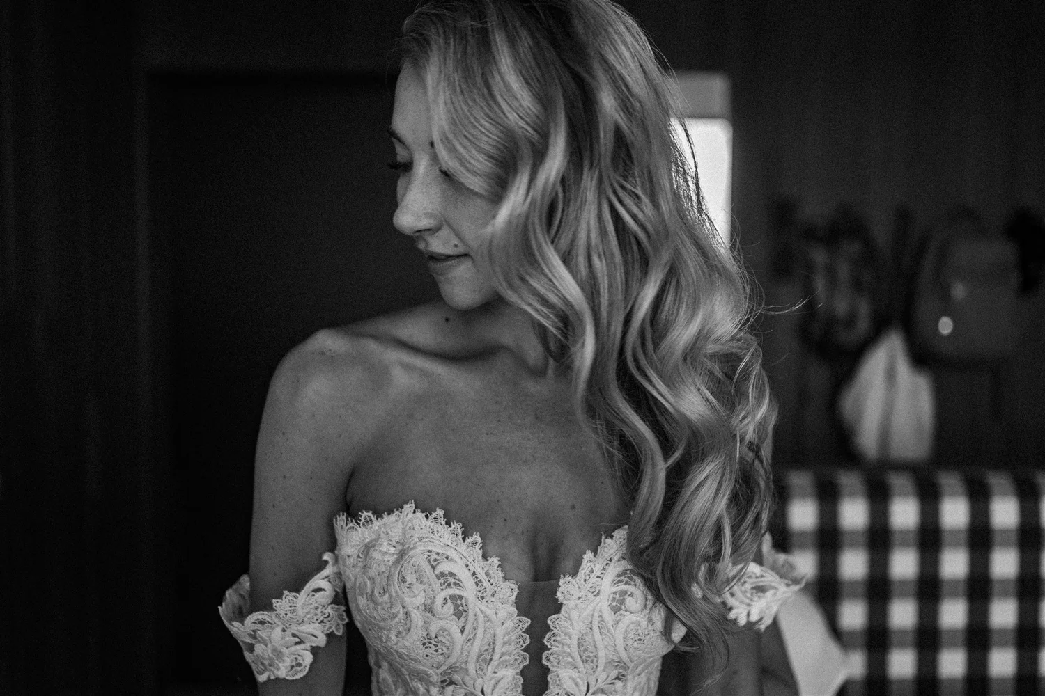 Black and white photo of a woman with long, wavy hair wearing lace dress, looking down and smiling, with a blurred figure in the background.