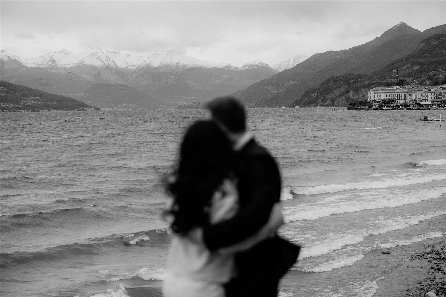 A blurred person standing on a beach near water with mountains in the background, some buildings along the shoreline, and cloudy skies.