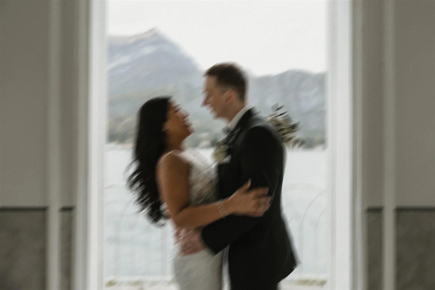 A blurry photo of a couple dressed in wedding attire, the woman in a white dress and the man in a suit, sharing a close moment with hills in the background.