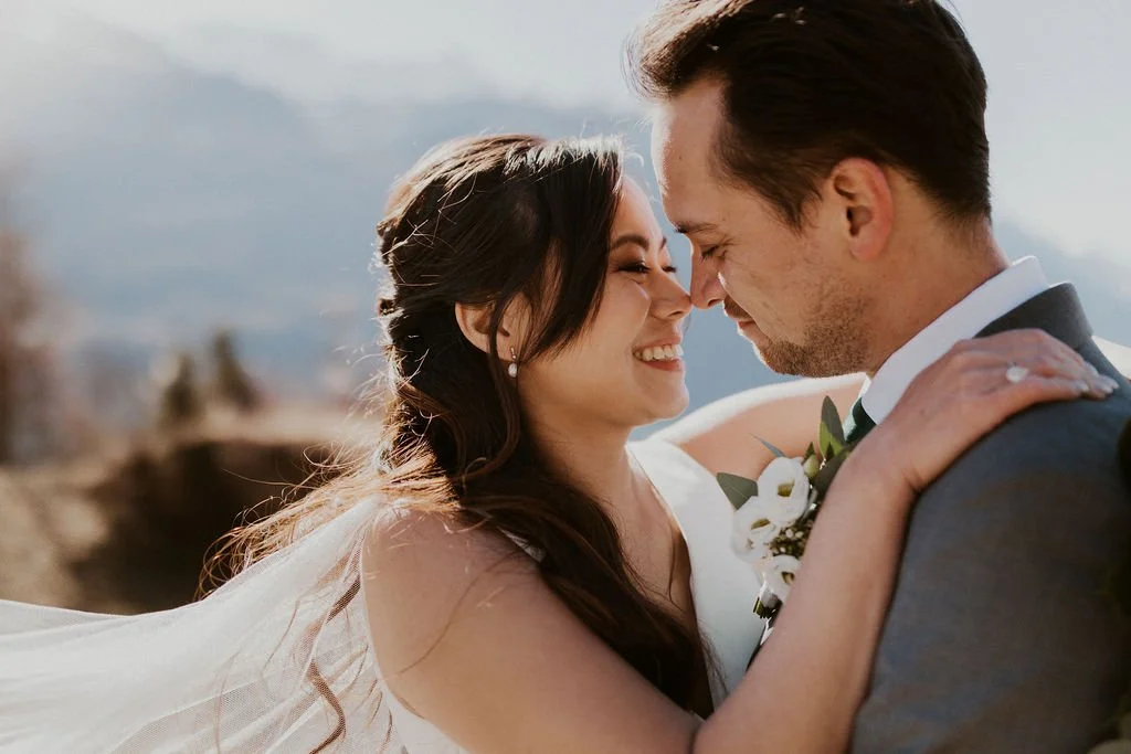 A bride and groom face-to-face, smiling with noses touching, outdoors with mountains in the background.
