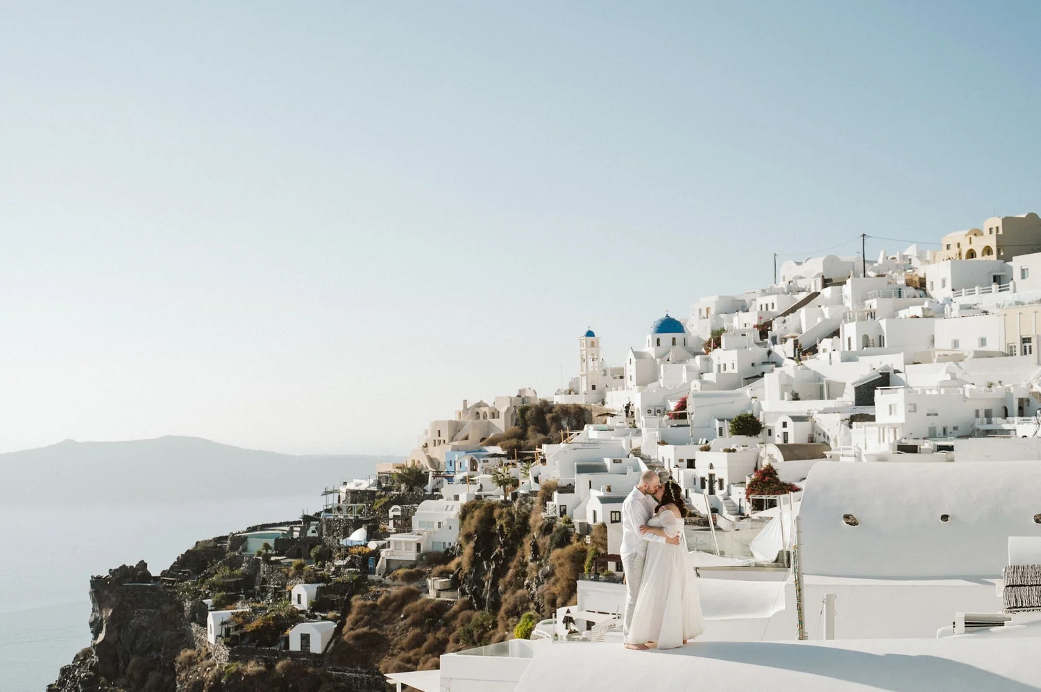 A couple, dressed in white wedding attire, sharing a kiss on a terrace overlooking white buildings on a hillside in Santorini, Greece, with the sea in the background.