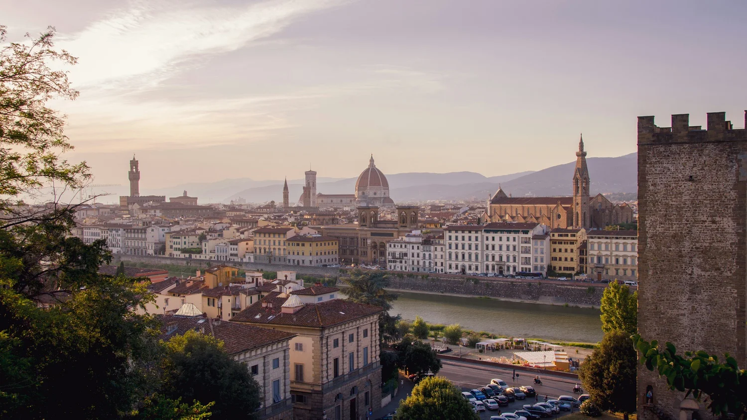Panoramic view of Florence, Italy, featuring the Florence Cathedral with a large dome, historical buildings, a river, and distant mountains during sunset.