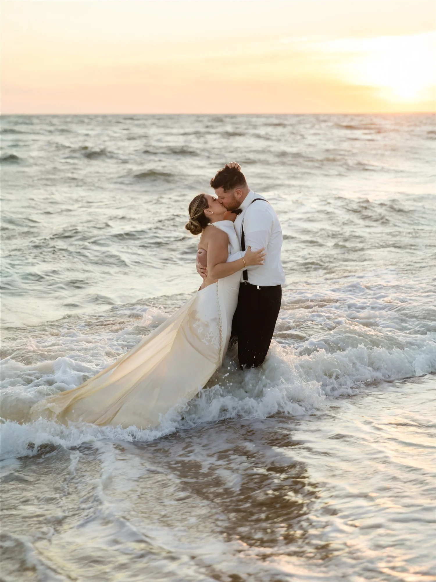 A newlywed couple in wedding attire sharing a kiss in the ocean at sunset.