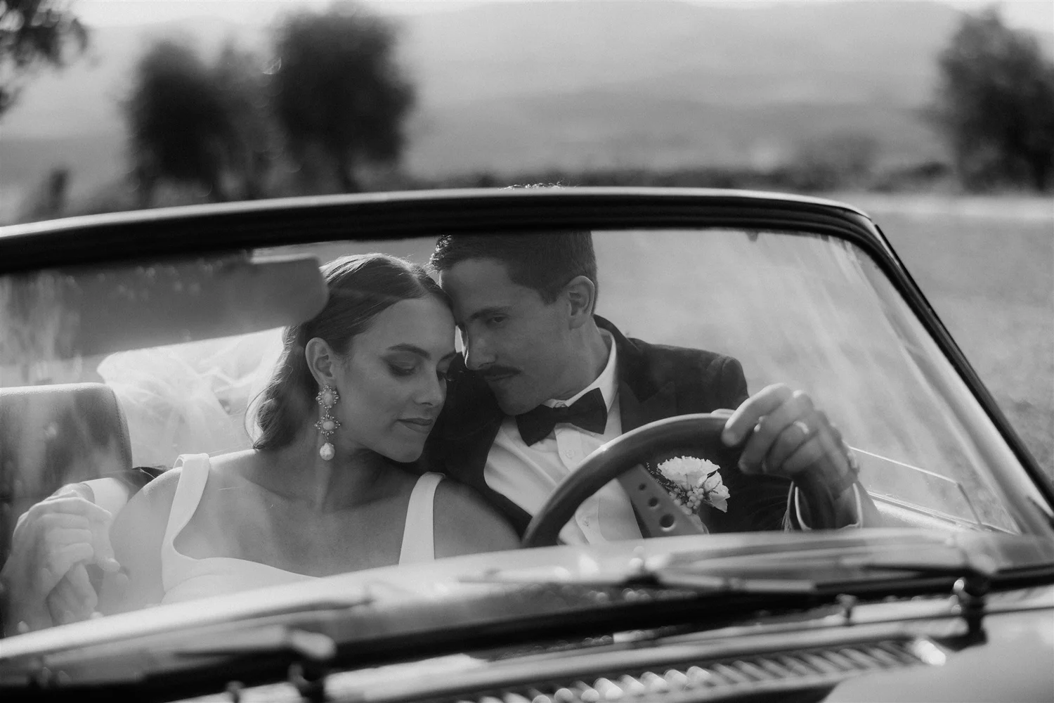 Black and white photo of a bride and groom sitting in a car, touching foreheads with eyes closed, appearing to be on their wedding day.