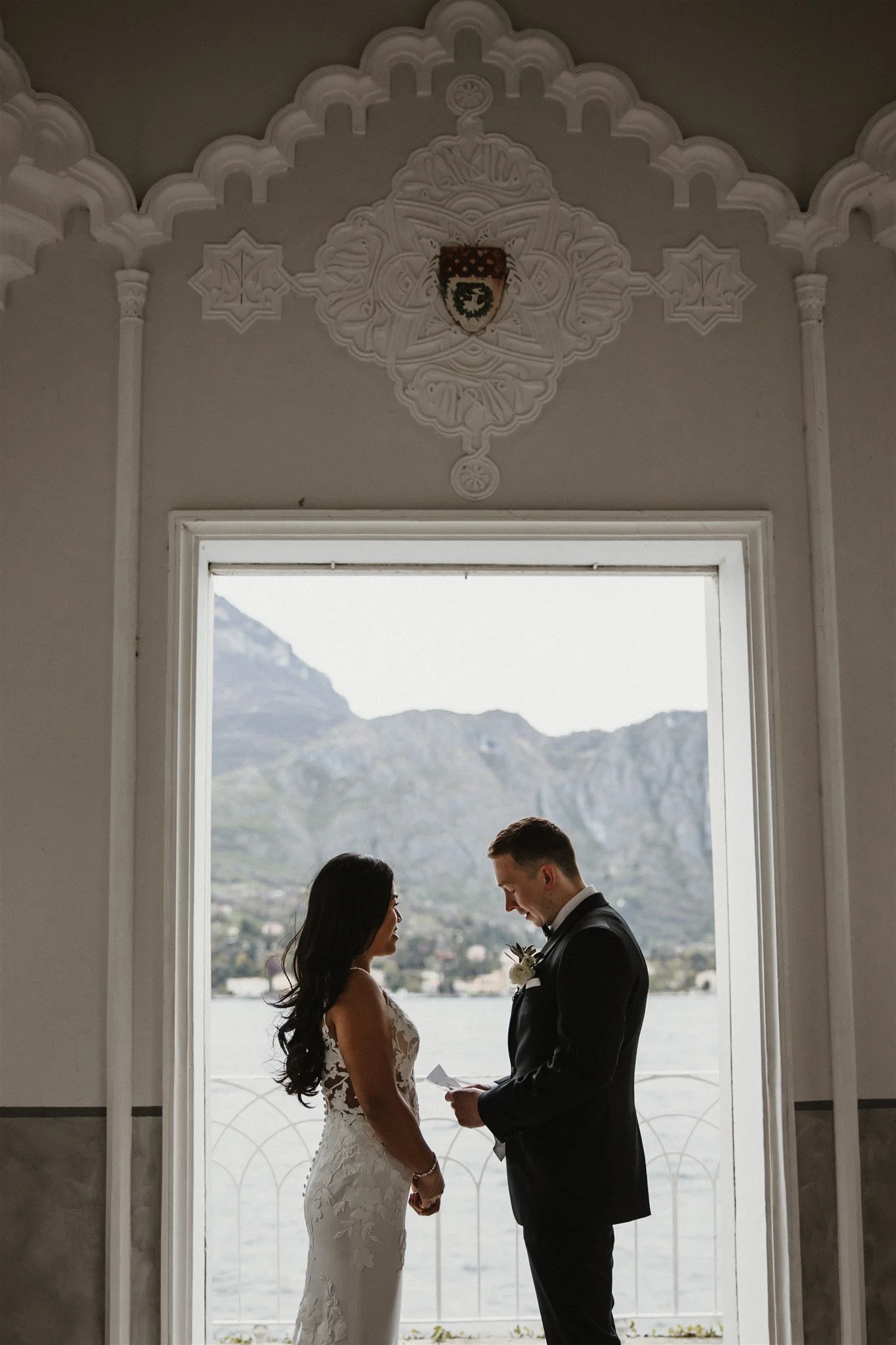 A bride and groom holding hands and facing each other in front of a large window with a scenic mountain and lake view, with decorative molding and a crest above the window.