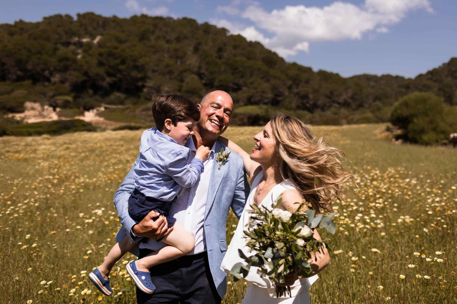 A family of three celebrating outdoors surrounded by a field of yellow flowers with green hills in the background. The father is carrying the young boy, both are smiling, and the woman is holding a bouquet of flowers.