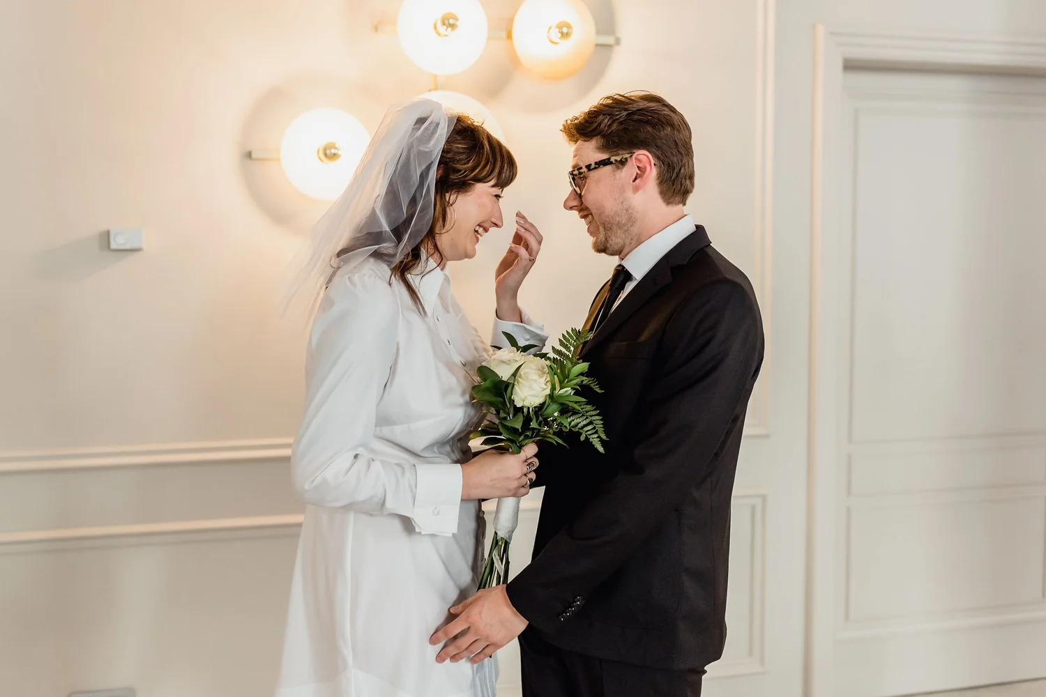 A bride and groom share a joyful moment during their wedding ceremony. The bride, wearing a white dress and veil, holds a bouquet of white roses and greenery. The groom, dressed in a black suit with glasses, smiles warmly toward her. They stand close