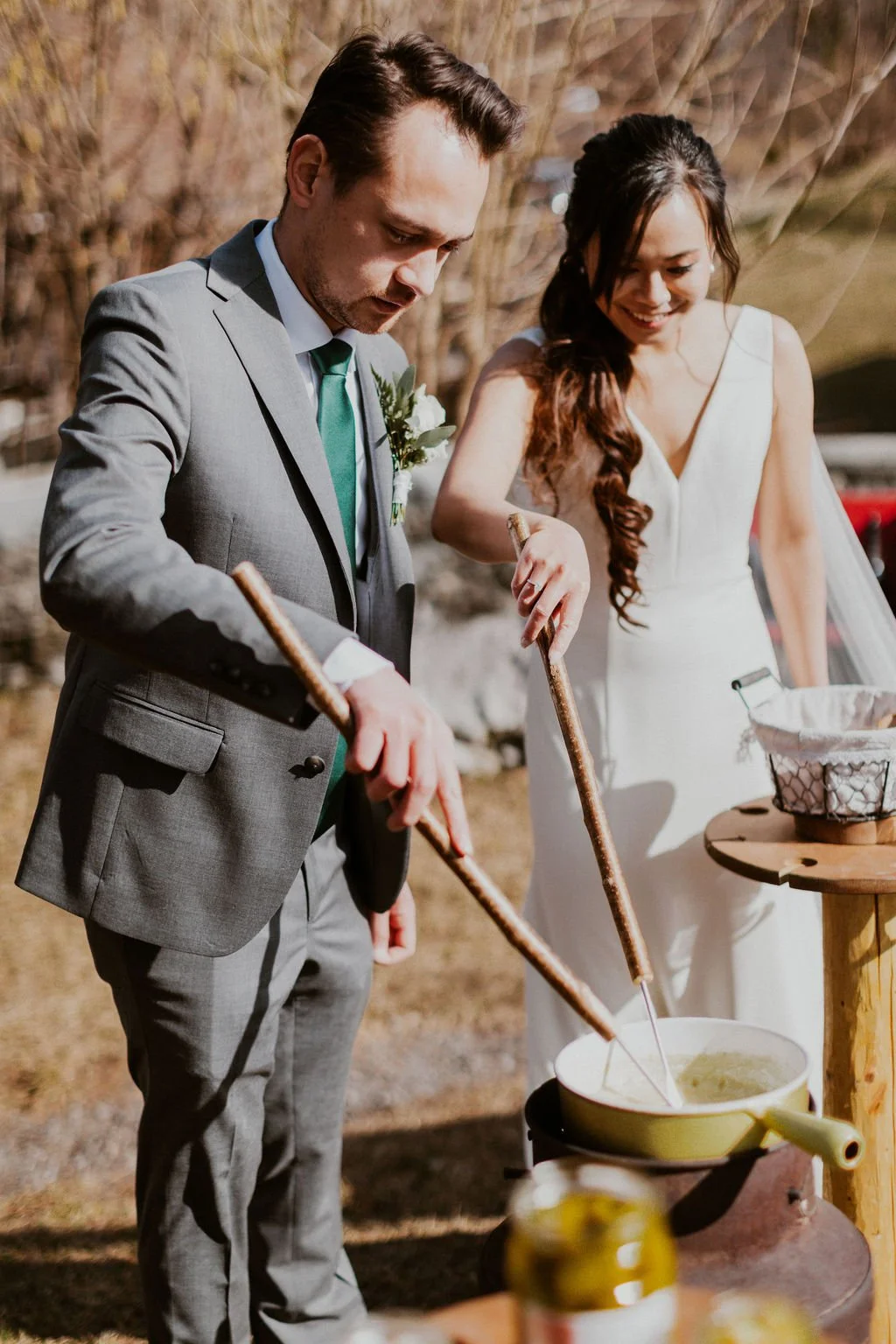 A bride and groom, dressed in wedding attire, are melting cheese over a fondue pot outdoors during daylight, smiling and enjoying their special moment.