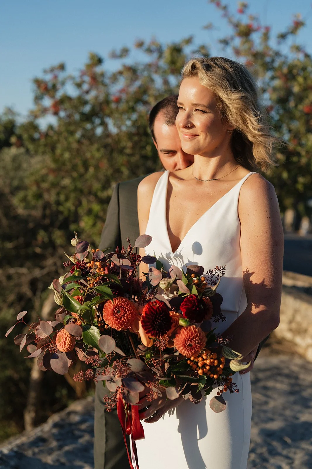 A woman in a white dress holding a colorful bouquet of flowers with a man standing behind her, outdoors with trees and clear sky in the background.