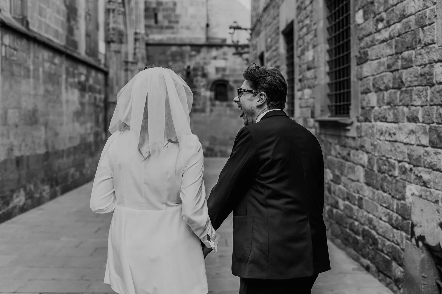 A black and white photo of a bride and groom holding hands, walking down an alleyway with brick walls on either side. The bride is wearing a white dress and veil, and the groom is wearing a dark suit with glasses. They are facing away from the camera