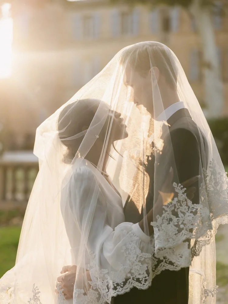 A bride and groom are standing close together outdoors, sharing a moment under a wedding veil on a sunny day.