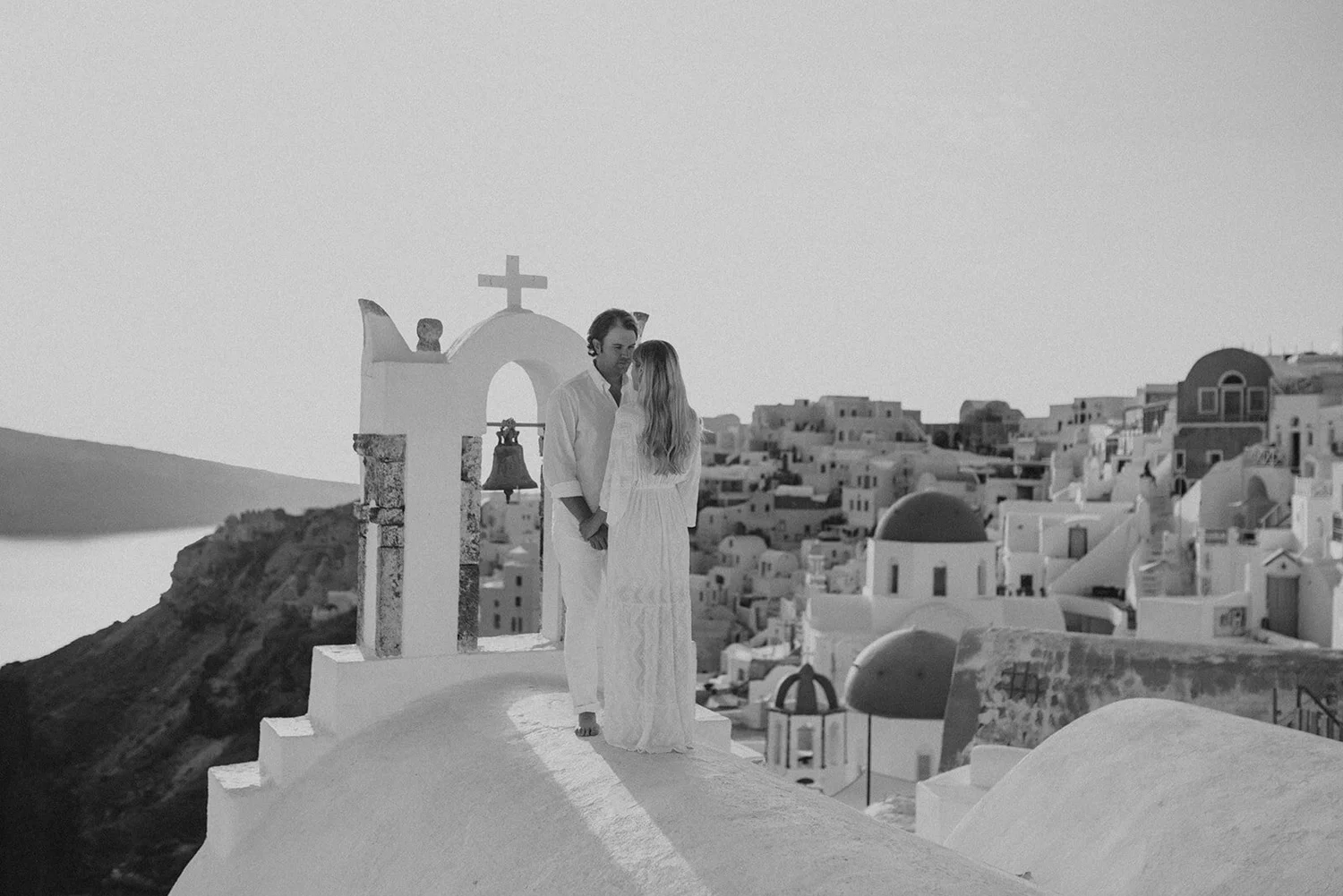 A black and white photo of a couple standing on a white rooftop ledge in Santorini, Greece, holding hands and gazing into each other's eyes with the island's white buildings and domed churches in the background.
