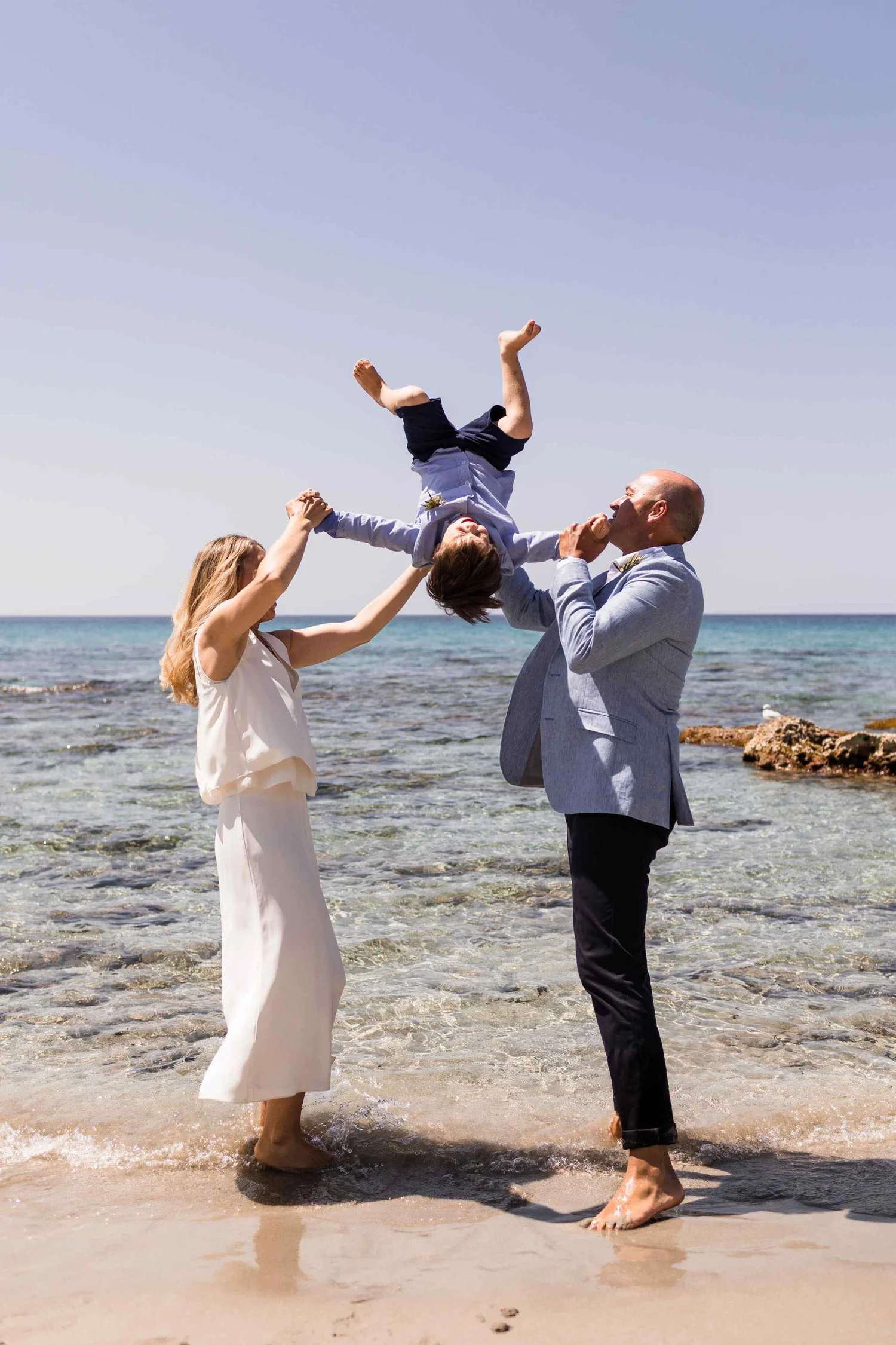 A family of three at the beach, with a woman and man holding a young boy upside down between them, all smiling. The woman has long blonde hair, wears a white dress, and is barefoot. The man is bald, wears a gray blazer with black pants, and is barefo