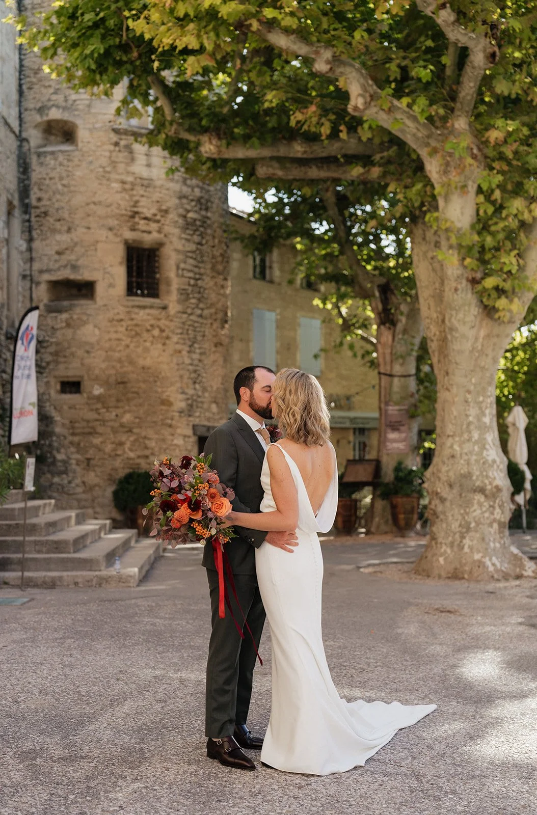 A newlywed couple sharing a kiss outdoors, the bride holding a bouquet of flowers, with an old stone building and large trees in the background.