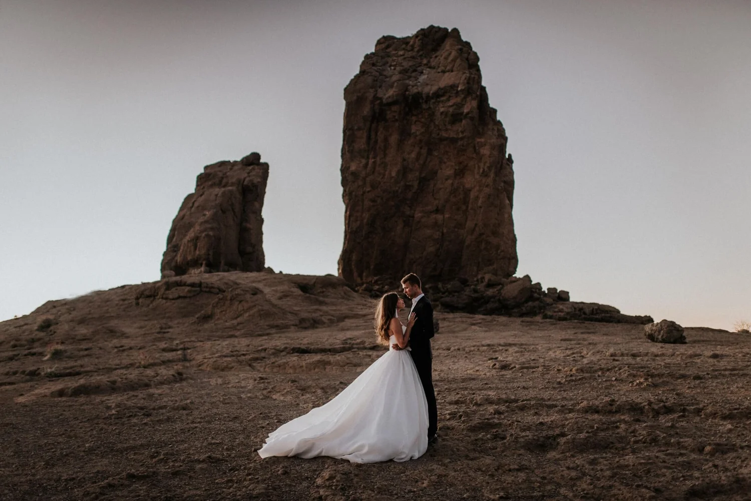 A bride and groom standing close together, facing each other, in a desert landscape. Behind them are two tall rock formations, with a cloudy sky overhead.