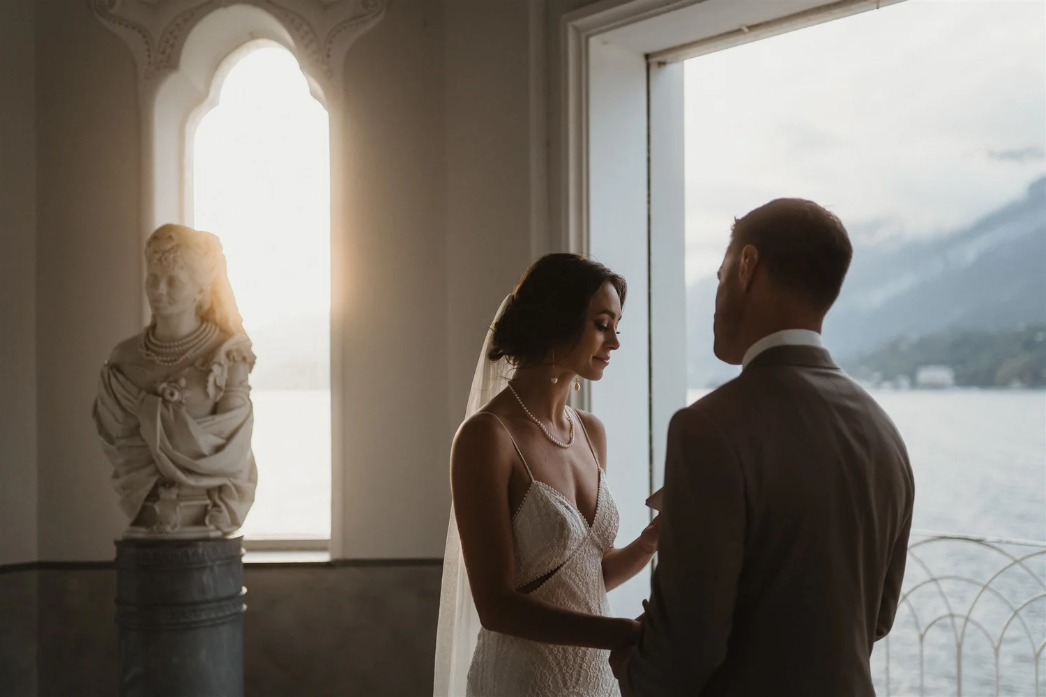 A bride and groom exchanging vows indoors with large windows and scenic view, with a classical bust statue on a pedestal nearby.