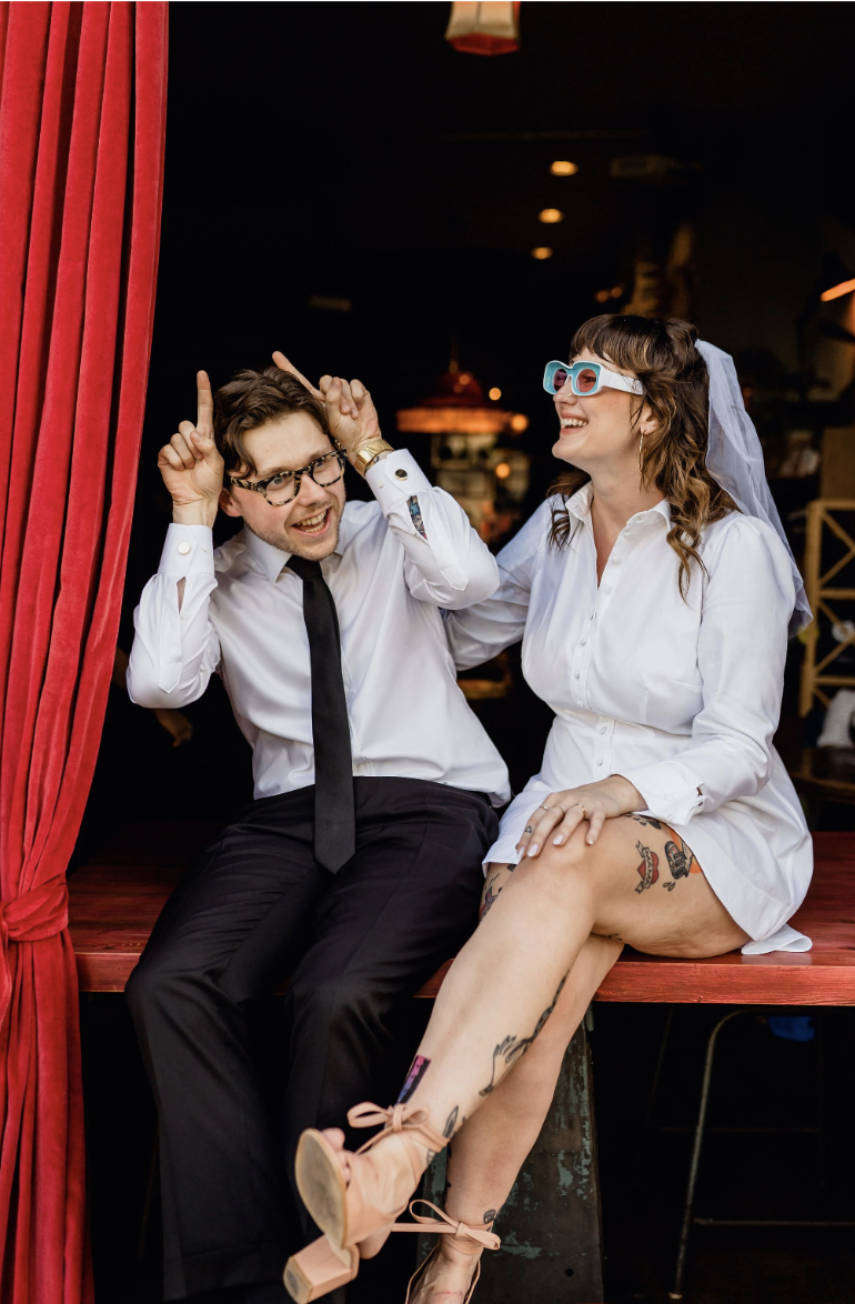 A man and woman sitting on a red stage with a red curtain to the side, both wearing white outfits. The man is wearing glasses, a white shirt, and a black tie, while the woman is wearing oversized 3D glasses, a white shirt, and has tattoos on her legs. They are sharing a joyful moment, laughing and making playful gestures.