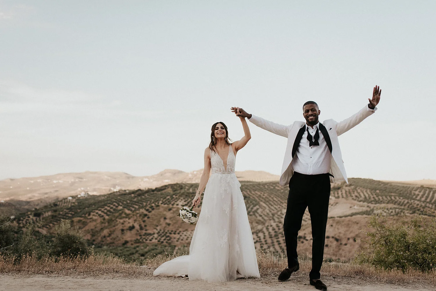 A couple in wedding attire holding hands and smiling outdoors in a hilly landscape.