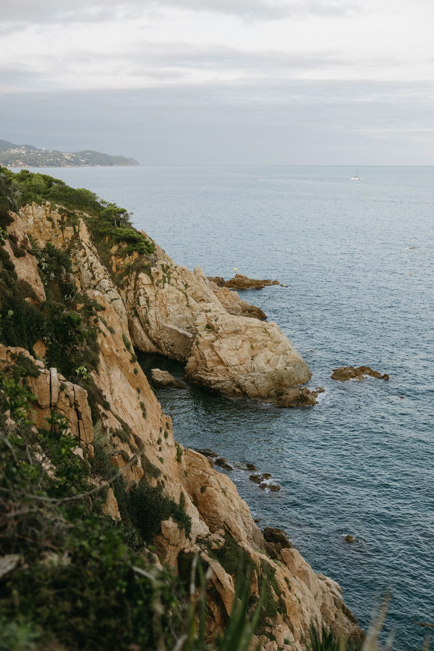 Cliffs and greenery along a rocky shoreline overlooking the ocean with a sailboat in the distance under cloudy skies.