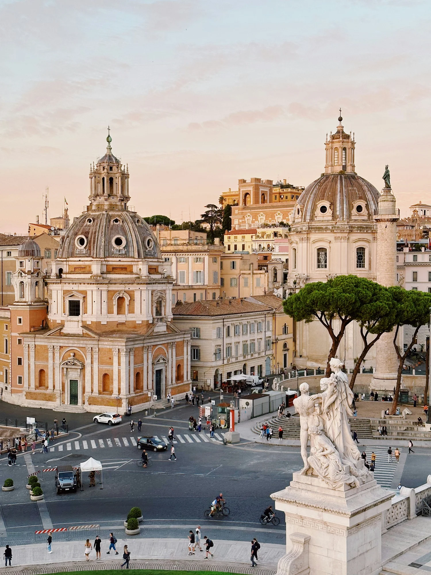 A view of Rome's historic city center at sunset featuring two domed churches, a street with cars and pedestrians, a statue of Roman gods, and a lively urban scene.