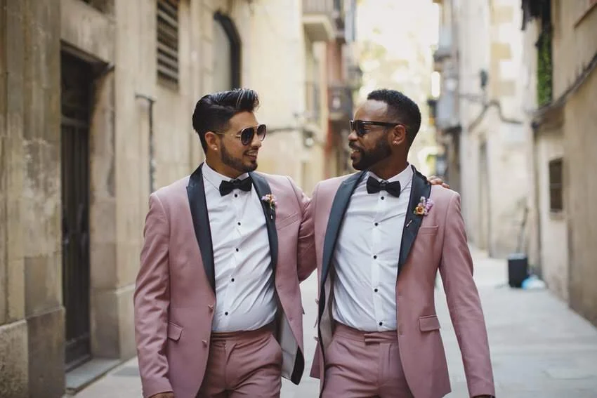 Two men in pink suits with black lapels, white shirts, and black bow ties walking arm in arm on a city street, smiling at each other.
