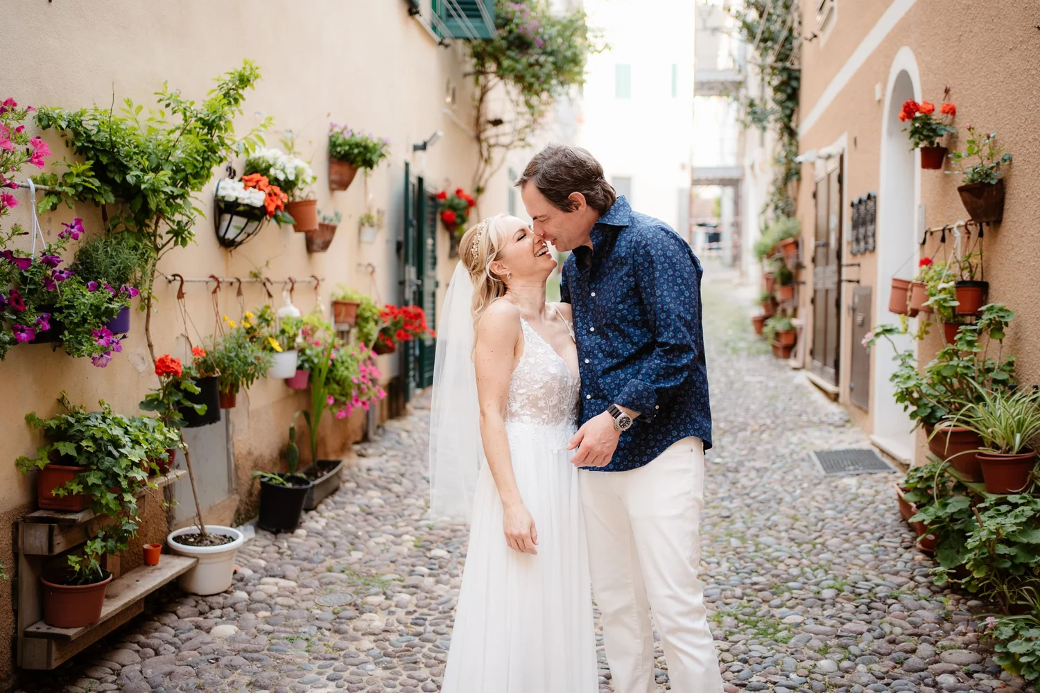 A couple, dressed in wedding attire, sharing a joyful moment on a cobblestone alleyway lined with colorful potted plants and flowers.