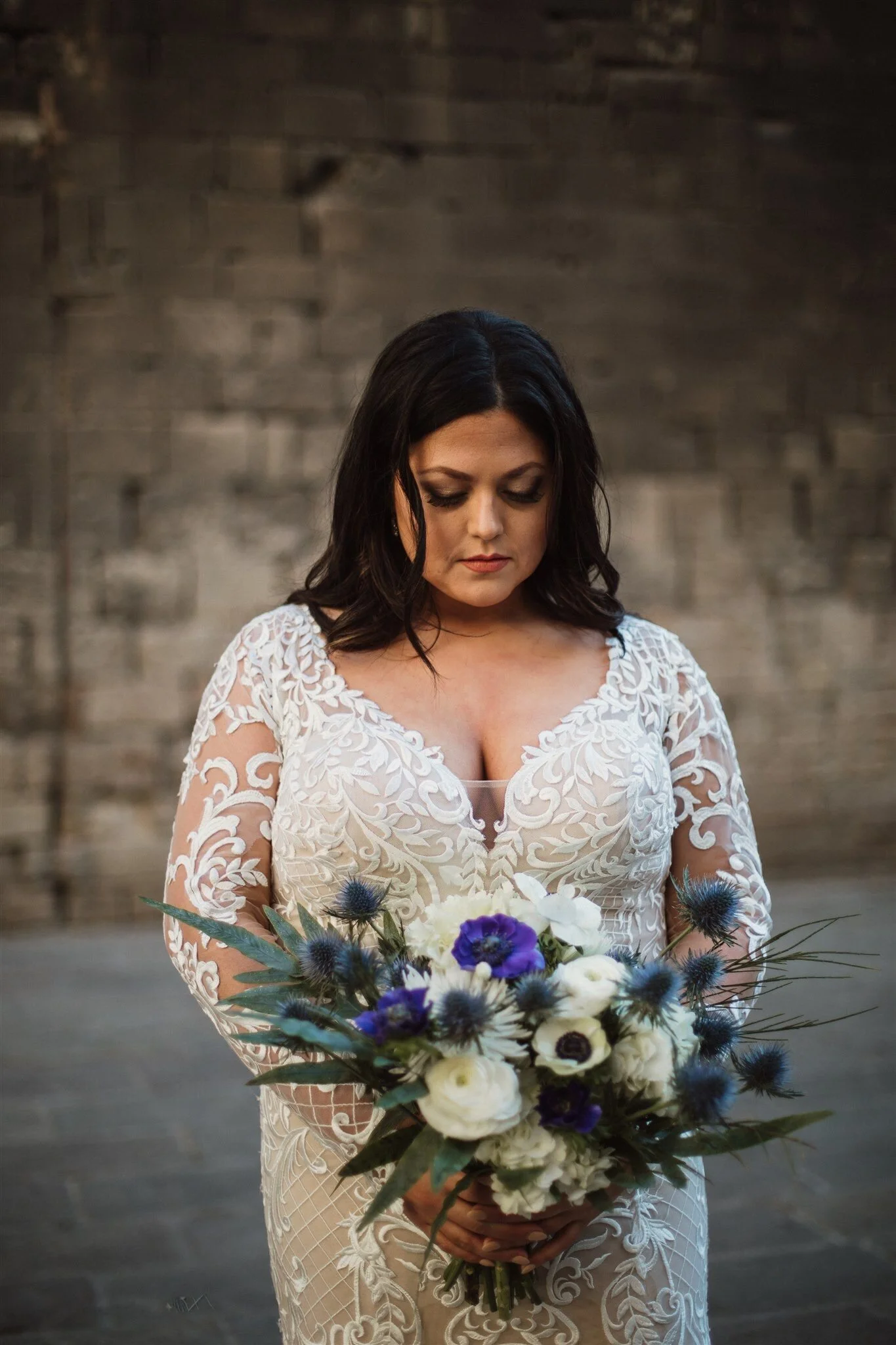 A woman in a white lace wedding dress holding a bouquet of white, purple, and blue flowers, standing against a dark brick wall with her head bowed, eyes closed.