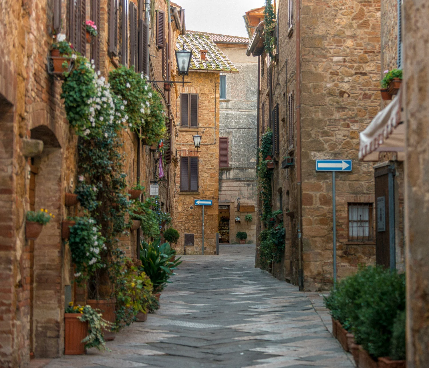 A narrow cobblestone alleyway lined with brick and stone buildings, featuring wooden window shutters, flower boxes, and street lamps. There are blue directional traffic signs pointing left and right along the alley.