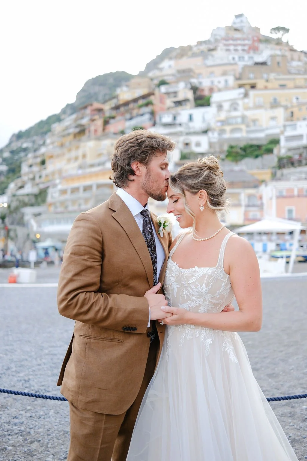 A bride and groom embrace in front of a hillside town, with the groom kissing the bride on her forehead as she smiles with her eyes closed.