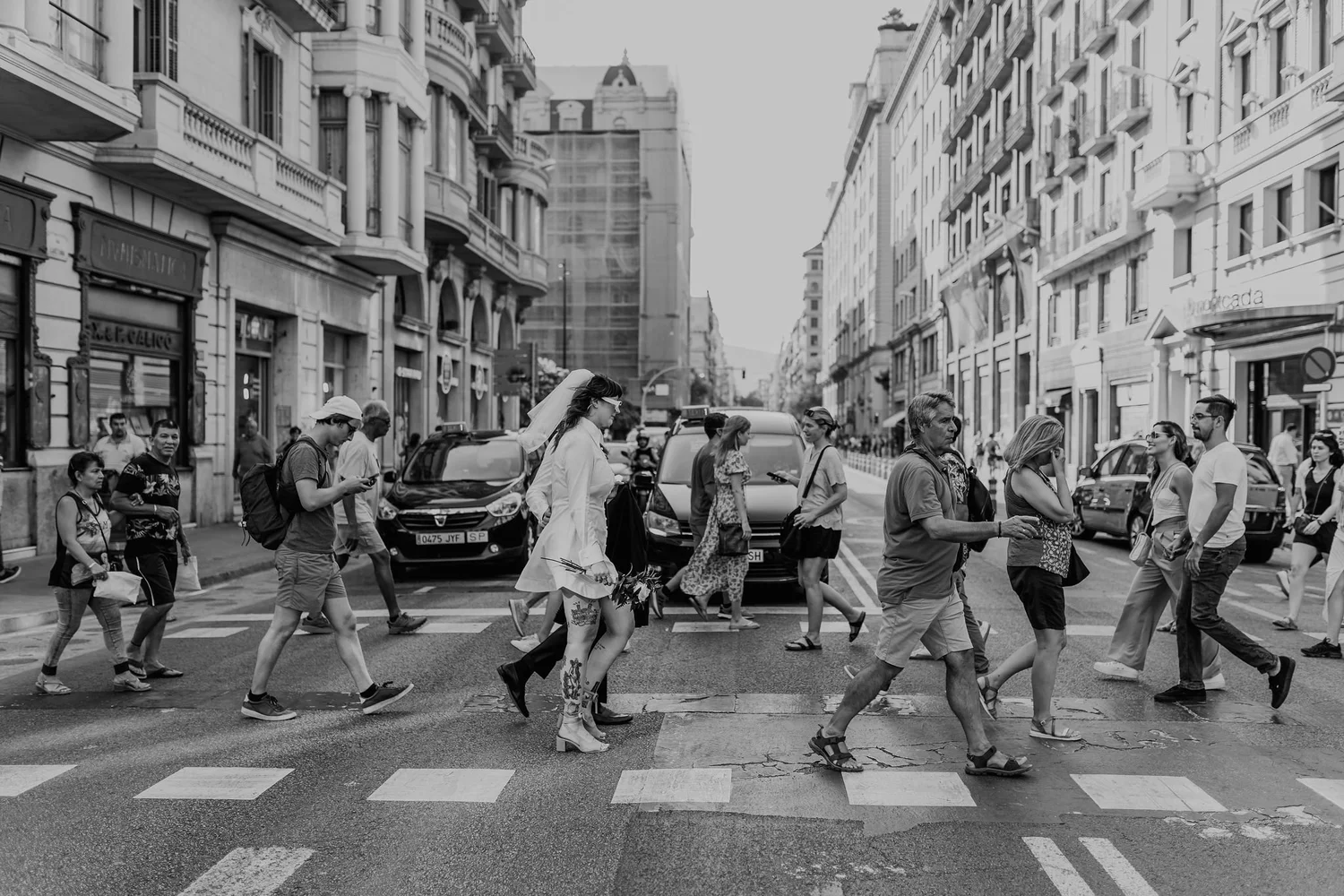 City street scene with pedestrians crossing at a crosswalk, surrounded by tall buildings with shops and apartments, black and white photograph.
