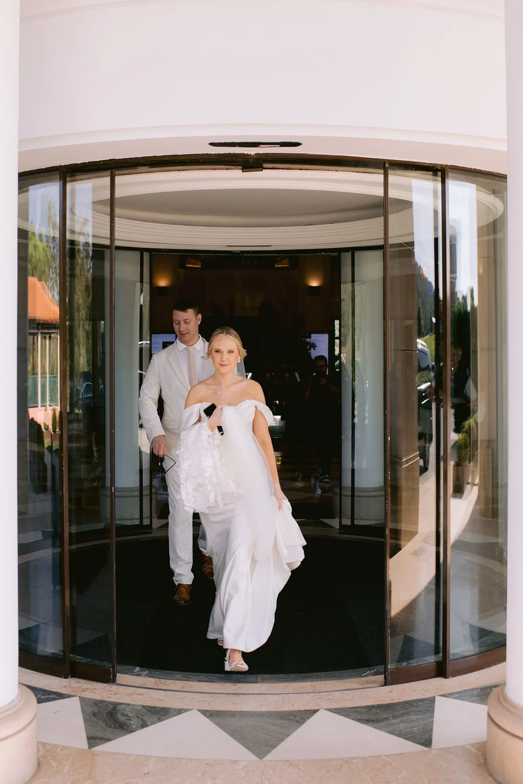 A bride in a white wedding dress walking out of a revolving glass door, accompanied by a man in a white suit, inside a building with white walls and decorative columns.