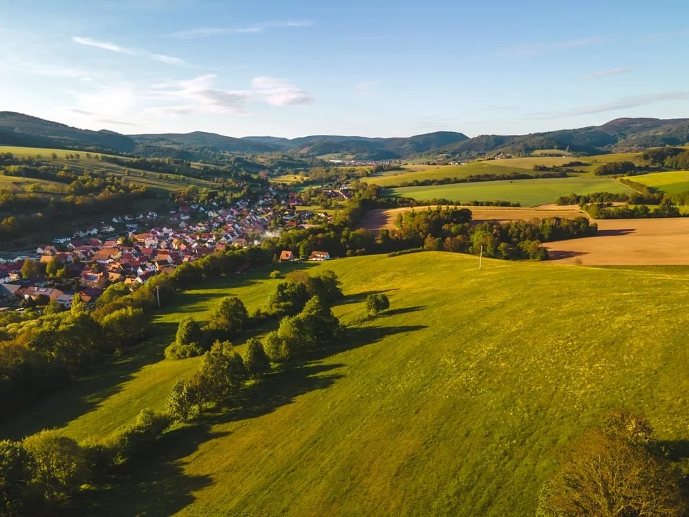 A scenic aerial view of a rural countryside with rolling green hills, a small town with houses, and distant mountains under a partly cloudy sky.