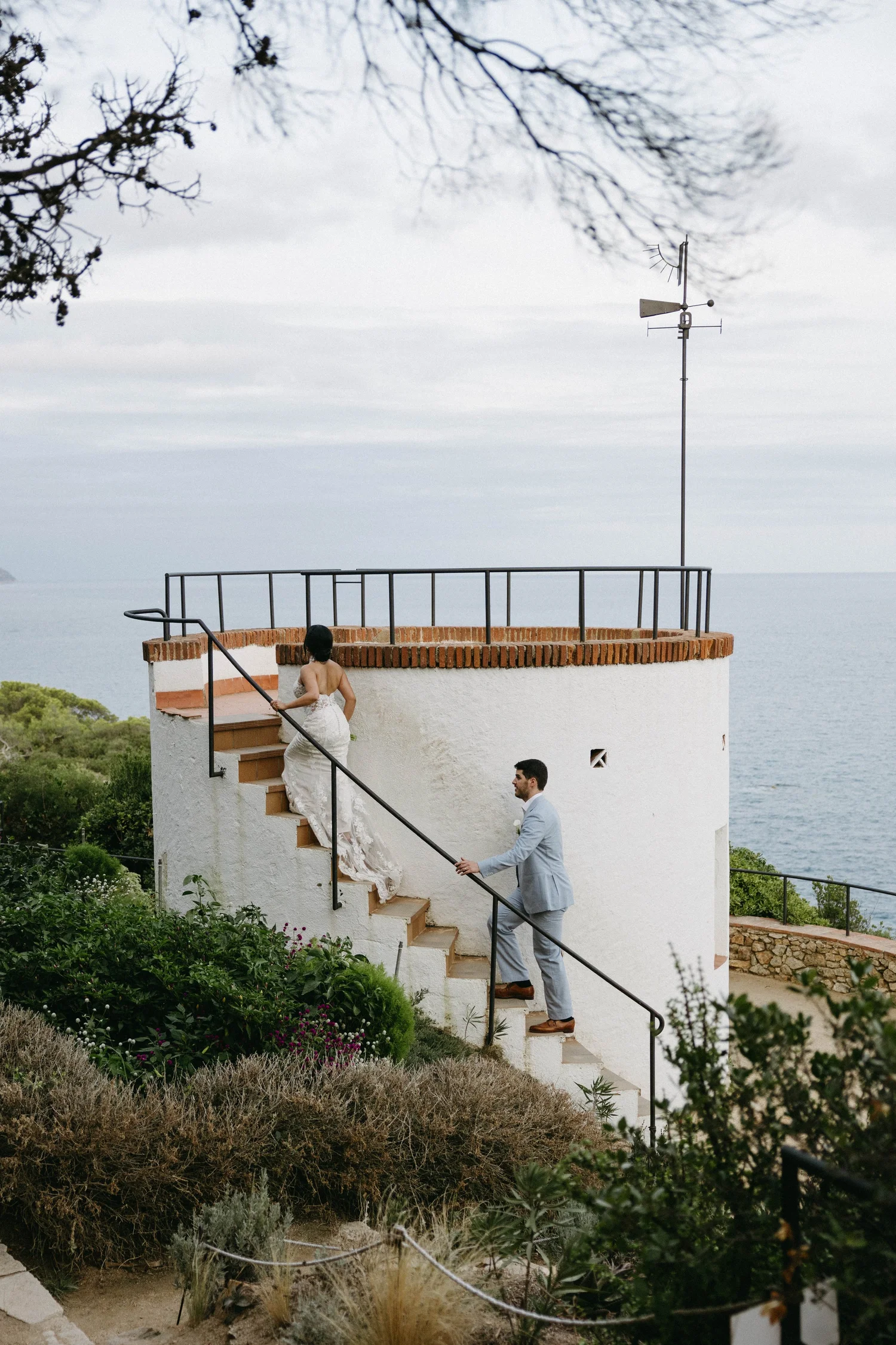 A bride in a white wedding dress is walking up stairs with a man in a light grey suit holding her hand, outdoors near the water, with greenery and shrubs in the foreground and a weather vane on the roof.
