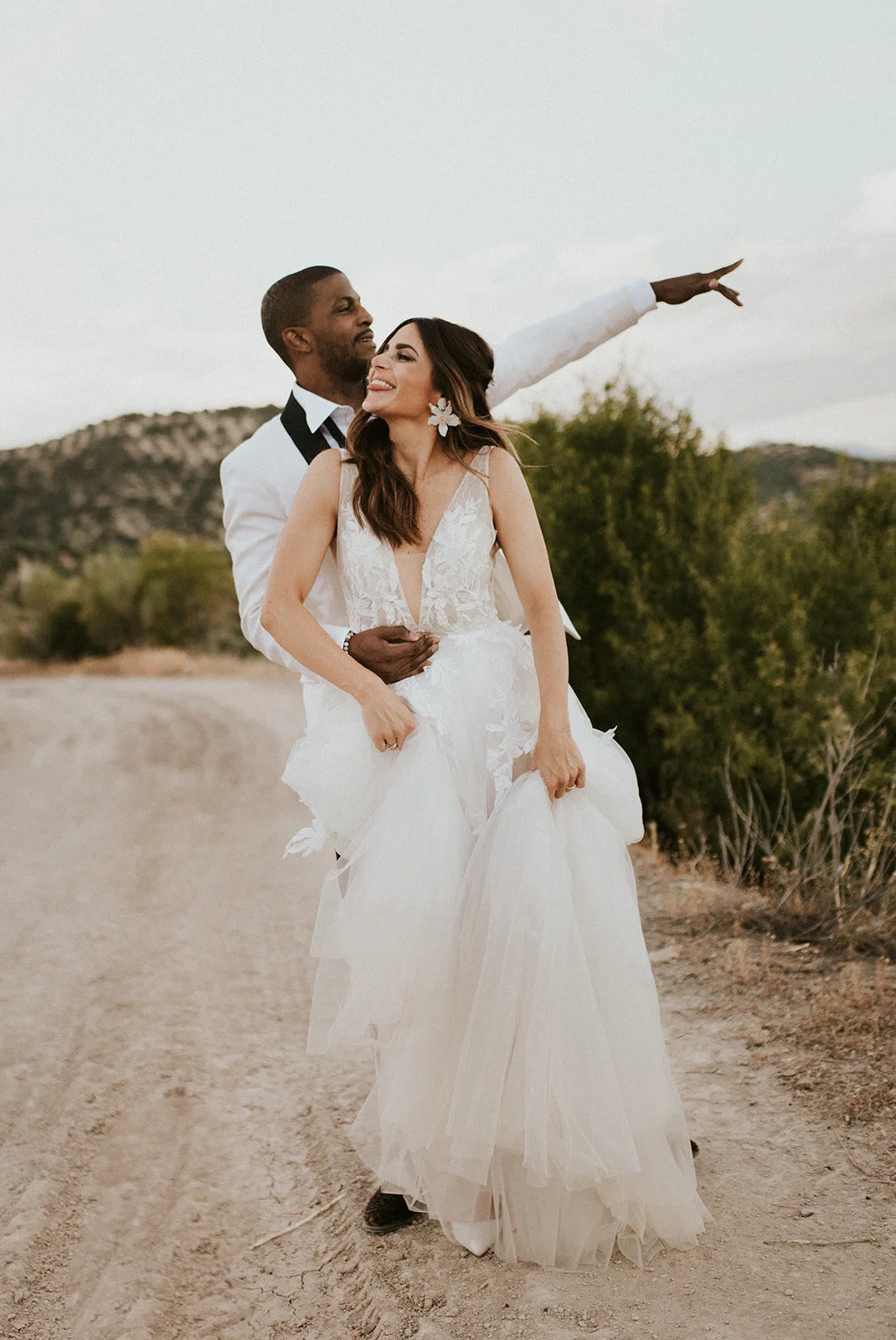 A newlywed couple standing on a dirt path outdoors, with the groom holding the bride and them smiling at each other, surrounded by natural scenery and hills in the background.
