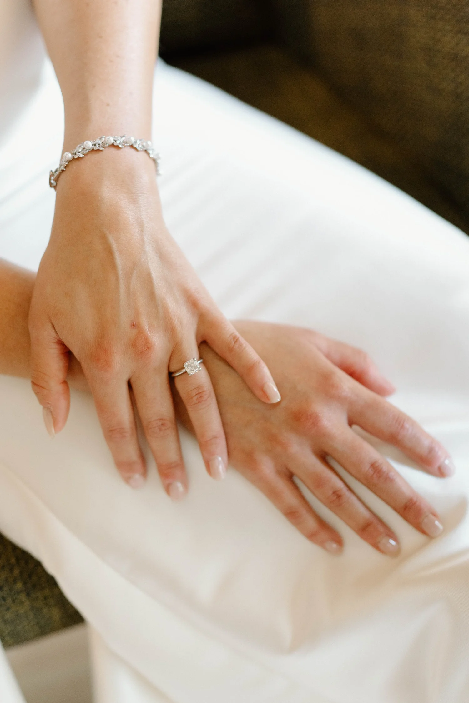 Close-up of two hands showing a wedding ring and bracelet on woman's hand, with a white fabric and brown background.