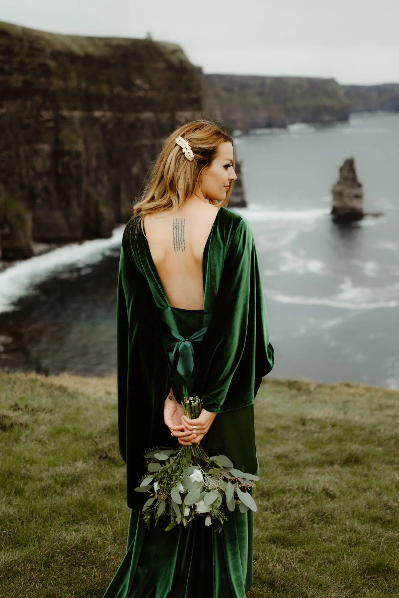 A woman with light brown hair styled with a hair clip, wearing a dark green velvet dress, standing by the ocean with cliffs in the background, holding a bouquet of greenery and white flowers.