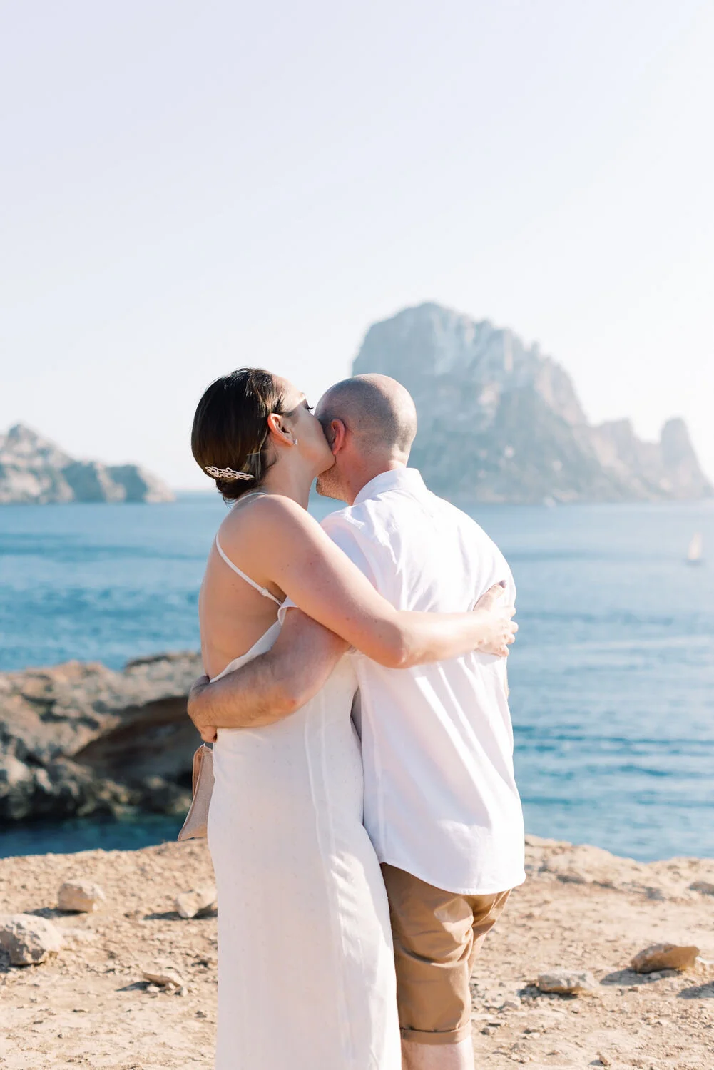 A couple embracing and sharing a kiss on a rocky beach with a large island and the ocean in the background.
