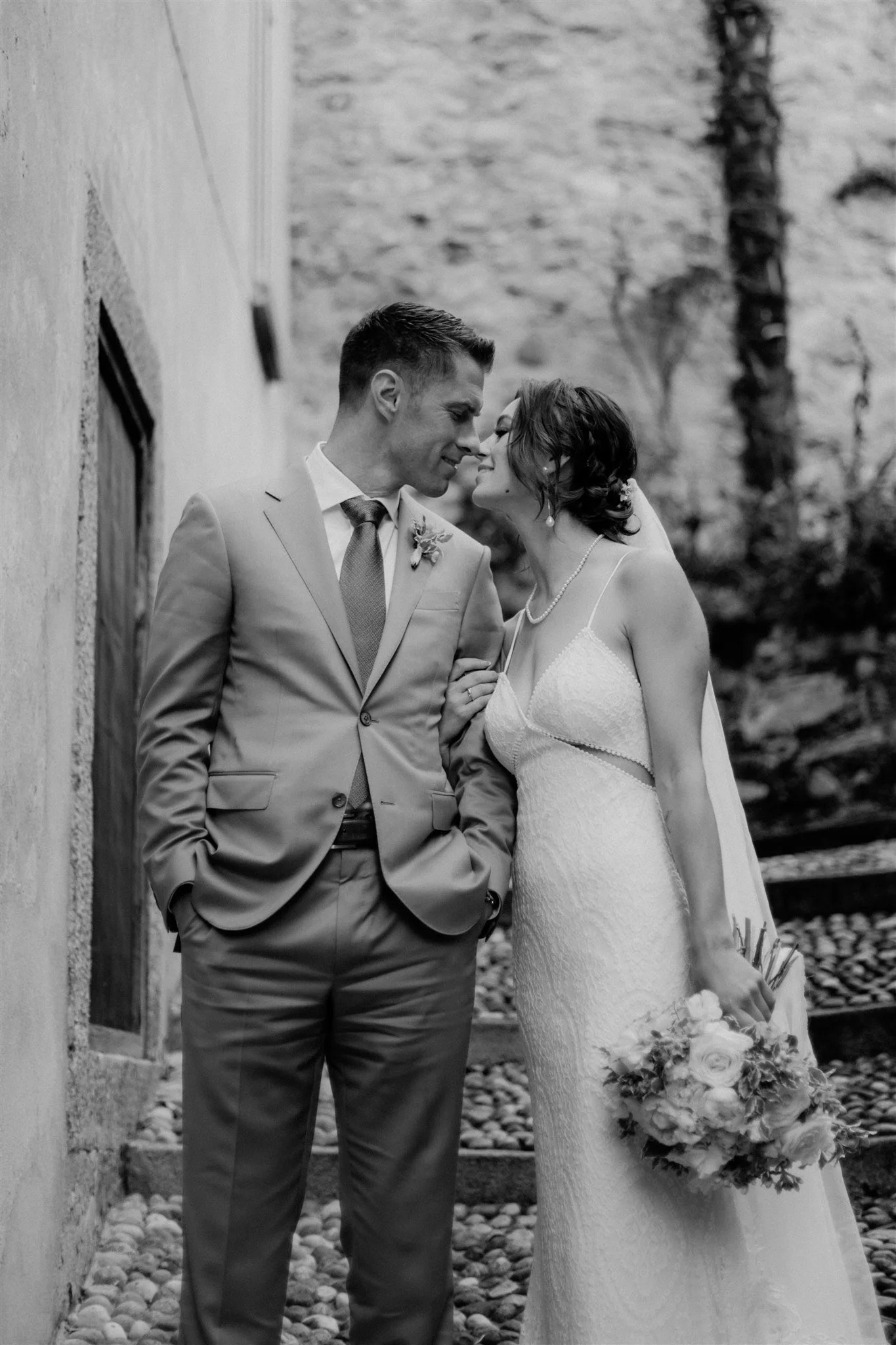 A black-and-white photo of a bride and groom facing each other, with foreheads touching in an outdoor setting. The groom is wearing a light-colored suit with a tie and boutonniere, and the bride is in a wedding dress holding a flower bouquet.