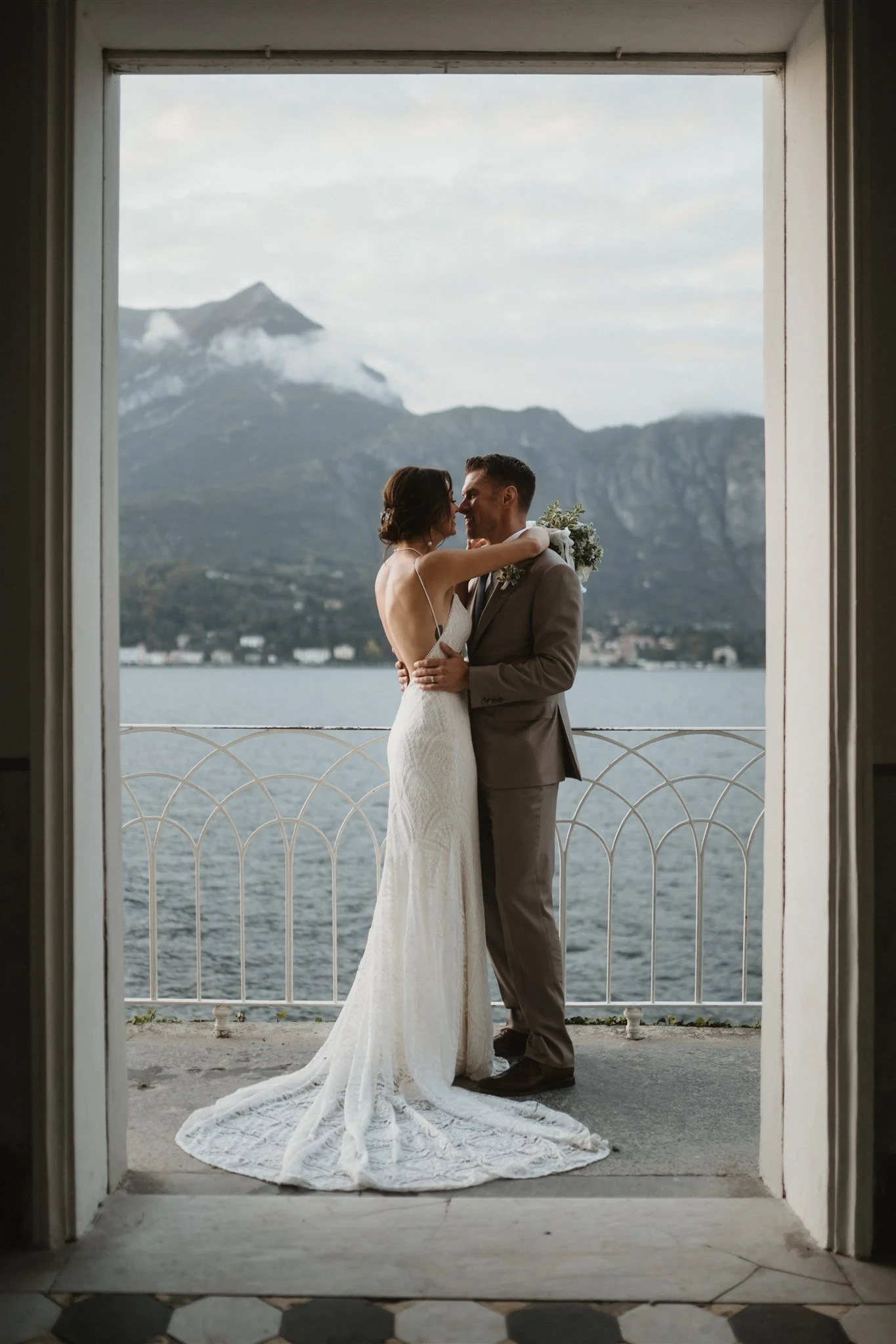 A bride and groom embrace on a balcony overlooking a lake and mountains, with a volcano in the distance.