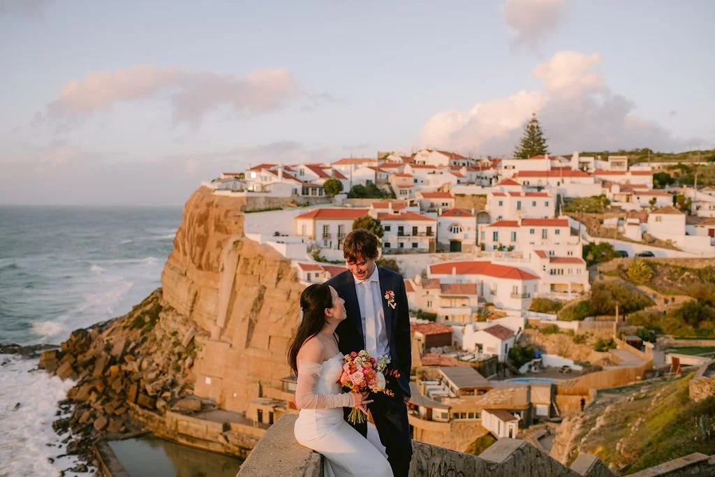 A couple in wedding attire sitting on a rock ledge overlooking a coastal village with white buildings and red roofs at sunset.