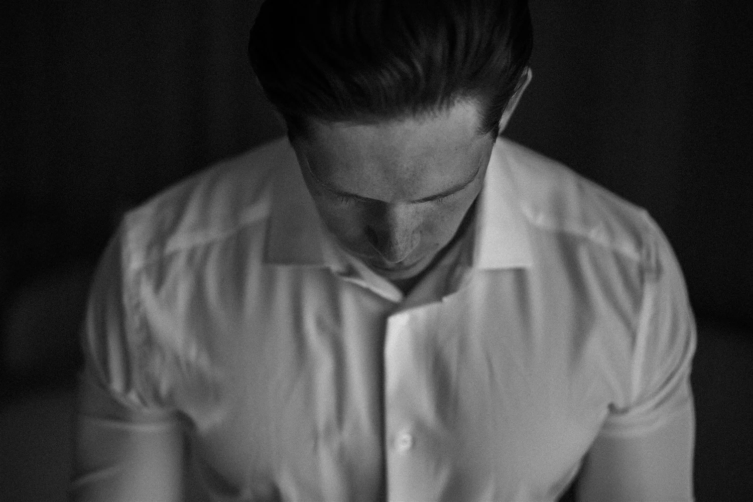 A monochrome photo of a man with slicked-back hair looking downward, wearing a collared shirt, and standing against a dark background.
