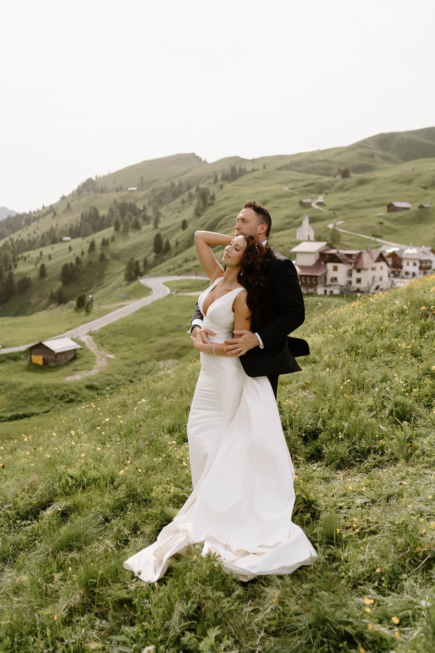 A bride and groom in wedding attire embracing outdoors on a grassy hillside with a winding road, small houses, and a church in a mountainous landscape.